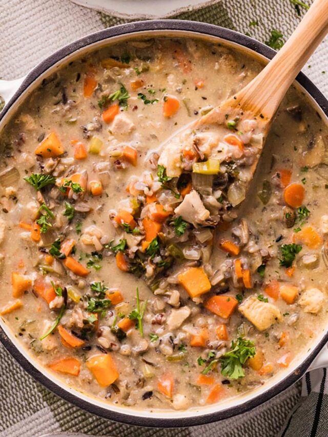Overhead of a Dutch oven filled with creamy chicken and wild rice soup—carrots, celery, mushrooms, and parsley—with a wooden spoon lifting a scoop.