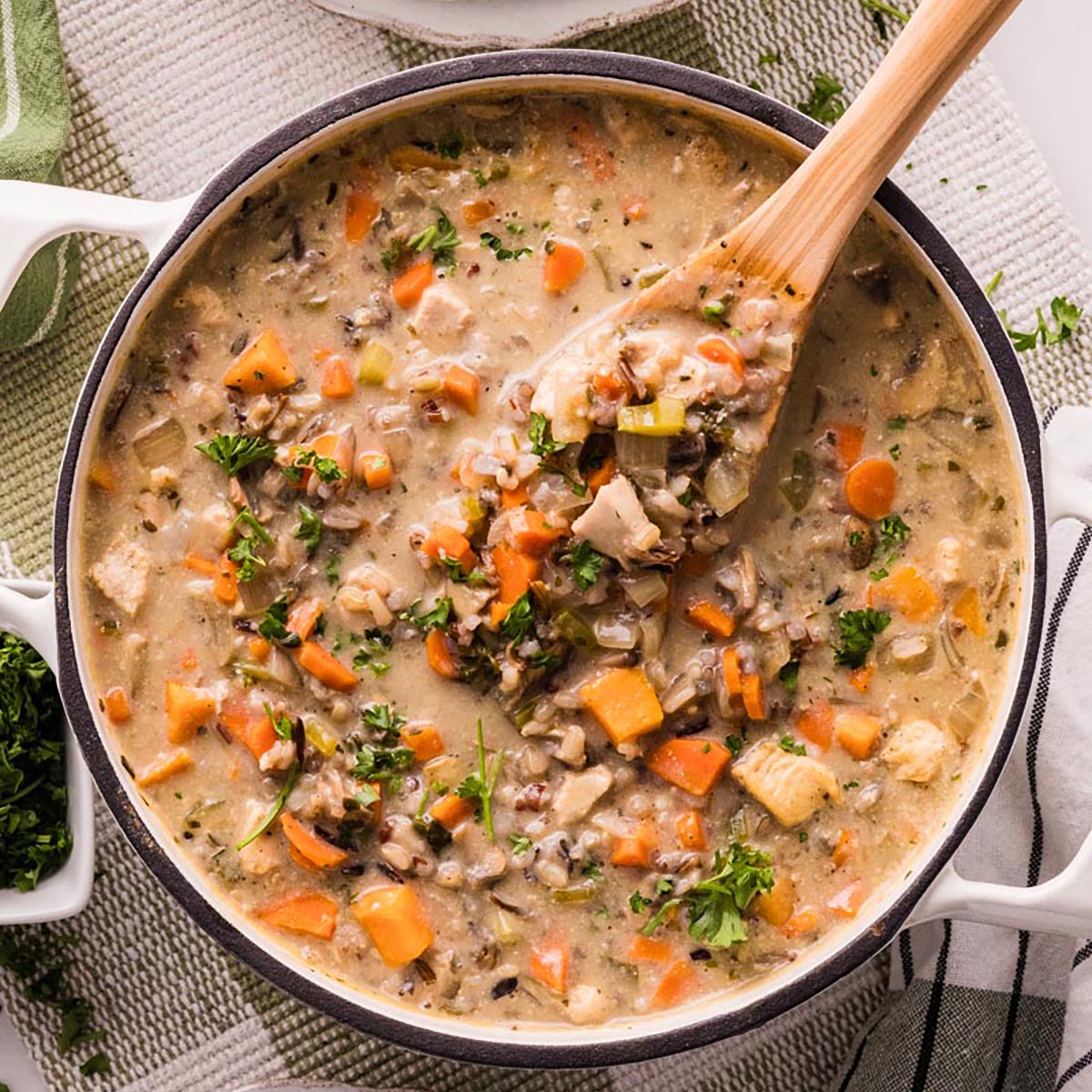 Overhead of a Dutch oven filled with creamy chicken and wild rice soup—carrots, celery, mushrooms, and parsley—with a wooden spoon lifting a scoop.