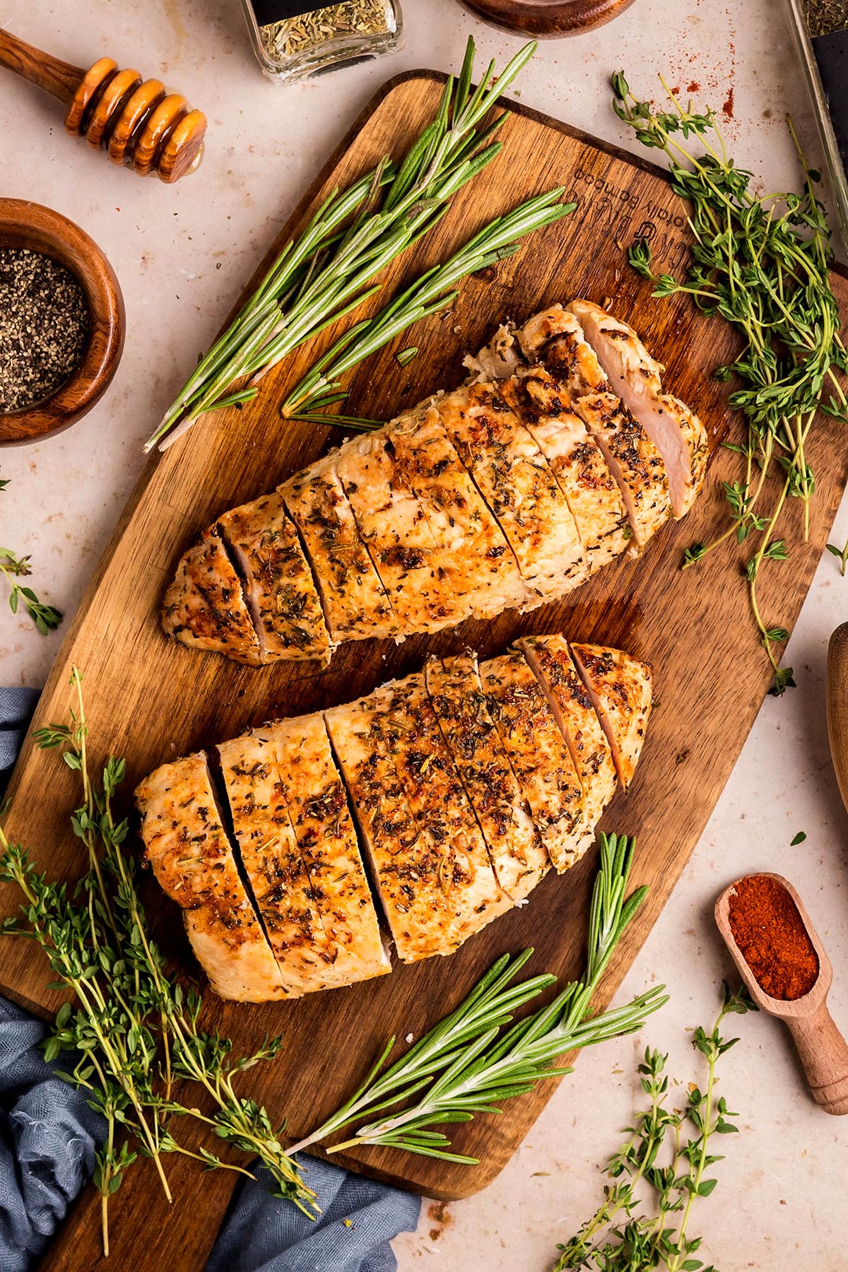 Overhead view of sliced oven-roasted turkey tenderloins on a wooden board with rosemary, thyme, and a honey dipper in the background.