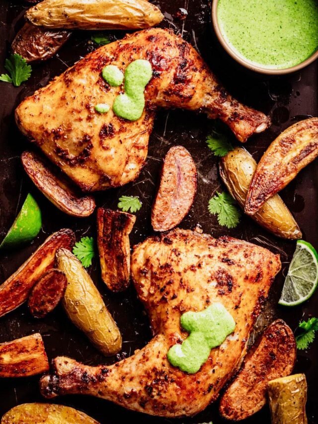 Sheet pan with Peruvian roasted chicken leg quarters topped with aji verde, crispy fingerling potatoes, cilantro leaves, lime wedges, and a small bowl of bright green sauce.