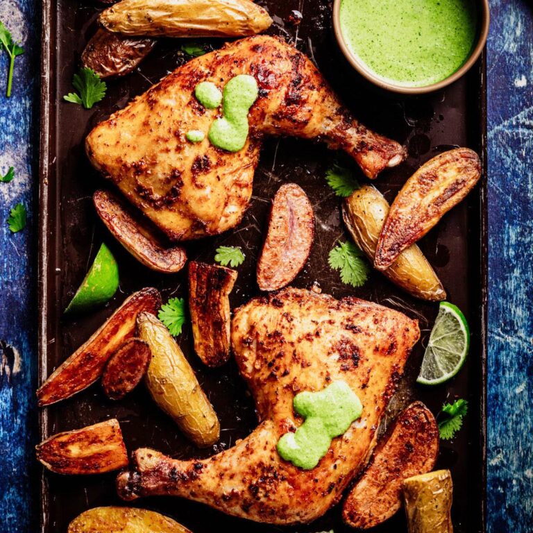 Sheet pan with Peruvian roasted chicken leg quarters topped with aji verde, crispy fingerling potatoes, cilantro leaves, lime wedges, and a small bowl of bright green sauce.