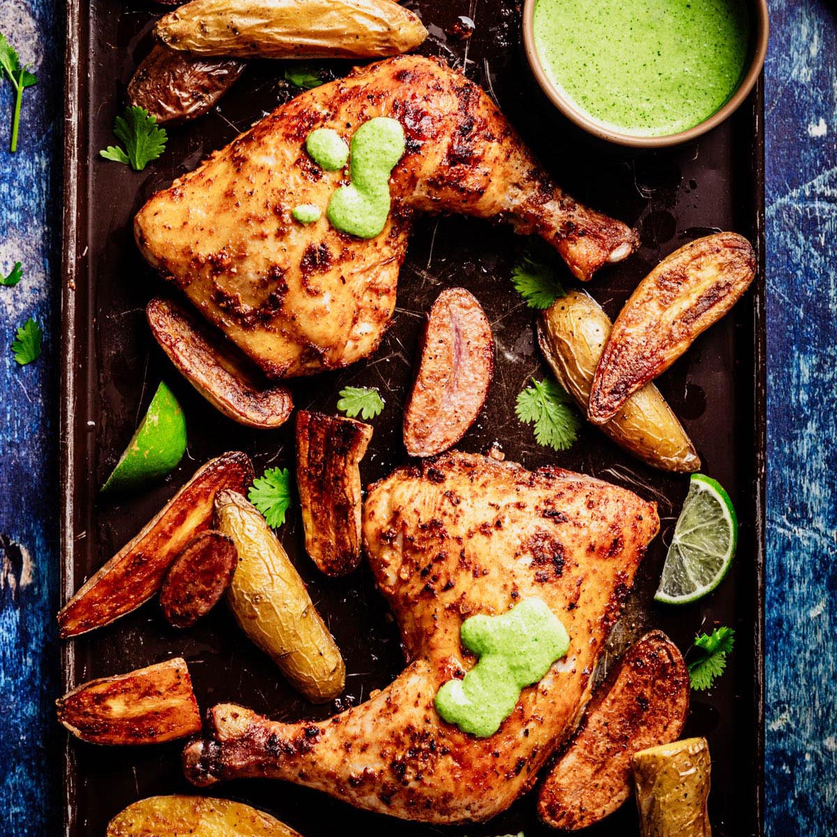 Sheet pan with Peruvian roasted chicken leg quarters topped with aji verde, crispy fingerling potatoes, cilantro leaves, lime wedges, and a small bowl of bright green sauce.