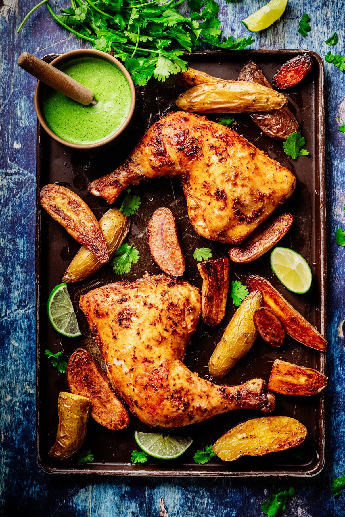 Sheet-pan Peruvian roasted chicken leg quarters with crispy fingerling potatoes, lime wedges, and cilantro, served with a bowl of bright green aji verde sauce on a blue surface.