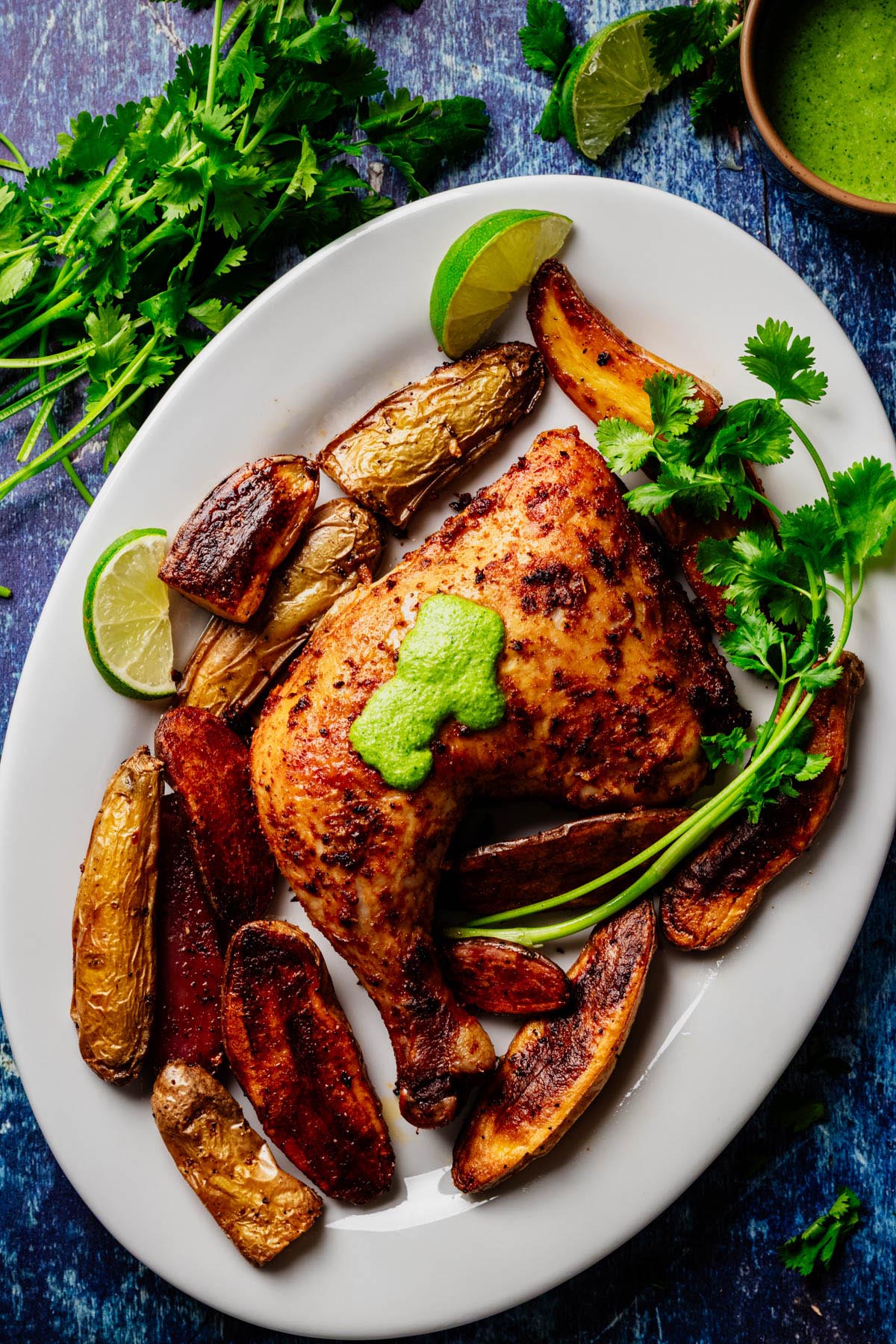 Plated Peruvian roasted chicken leg quarter topped with bright aji verde, surrounded by crispy fingerling potatoes, cilantro, and lime wedges on a blue tabletop.