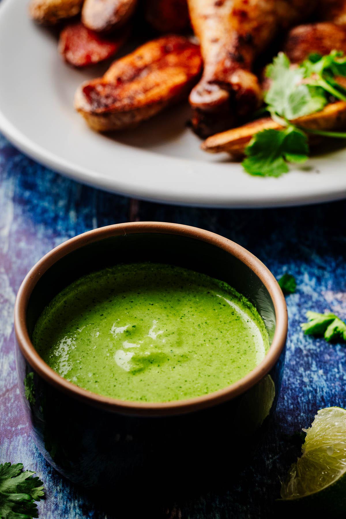 Small bowl of creamy green aji verde sauce with a plate of Peruvian chicken and roasted potatoes blurred in the background, cilantro and lime nearby.