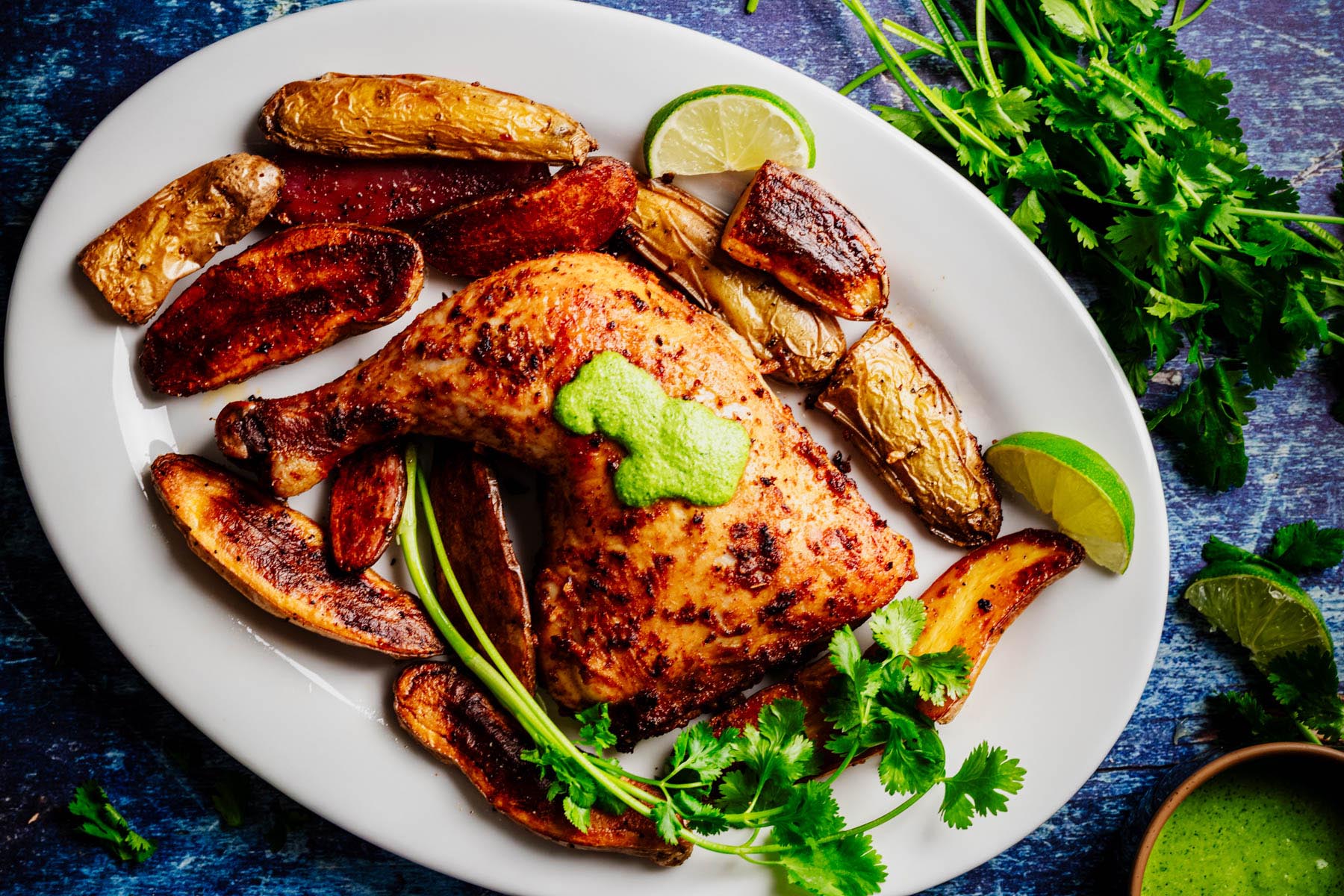 Peruvian roasted chicken leg quarter topped with aji verde, surrounded by crispy fingerling potatoes, cilantro, and lime wedges on a white platter.