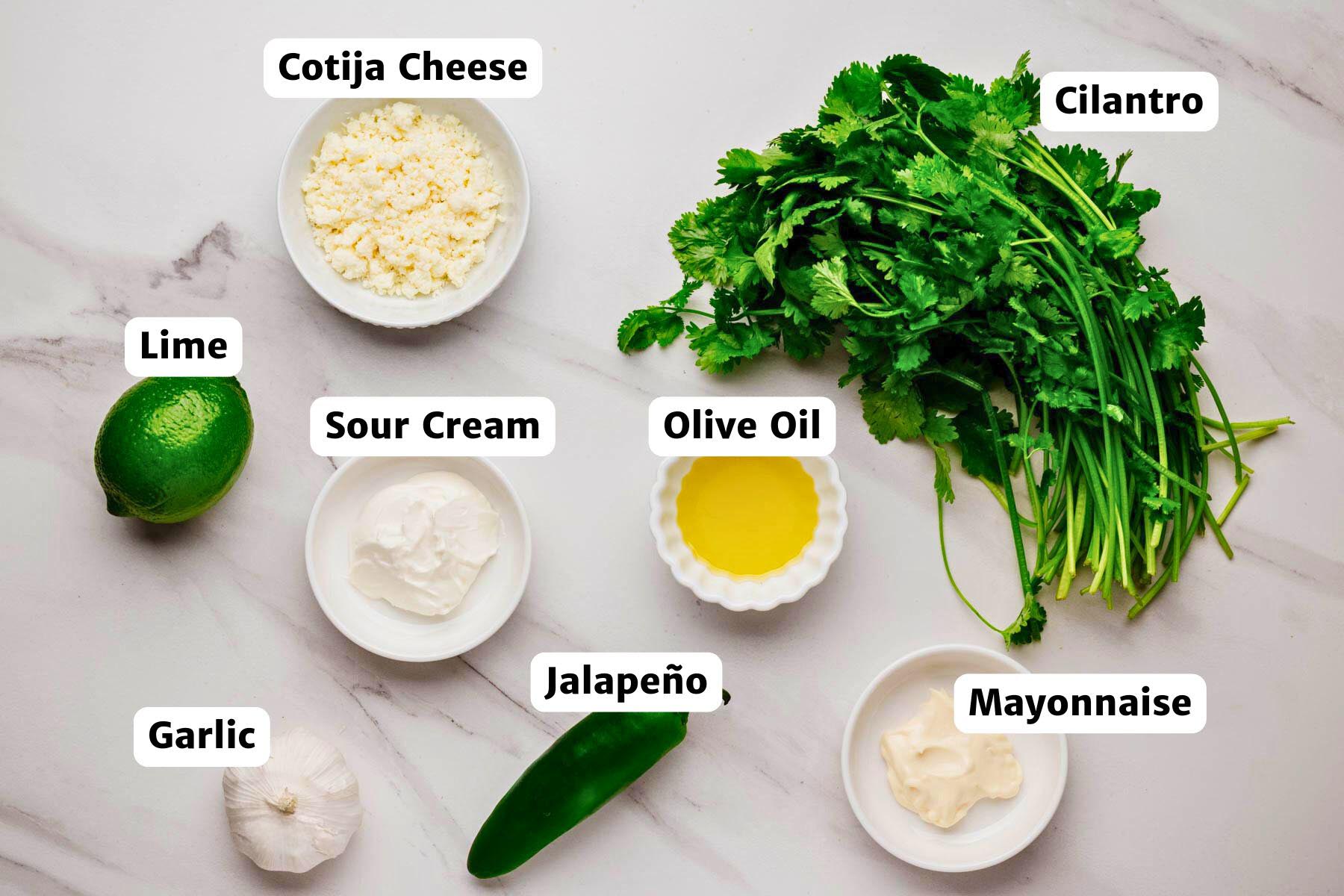 ngredients for aji verde sauce arranged on a marble surface—bunch of cilantro, bowl of crumbled cotija, lime, sour cream, olive oil, jalapeño, mayonnaise, and a garlic head—with labeled callouts.