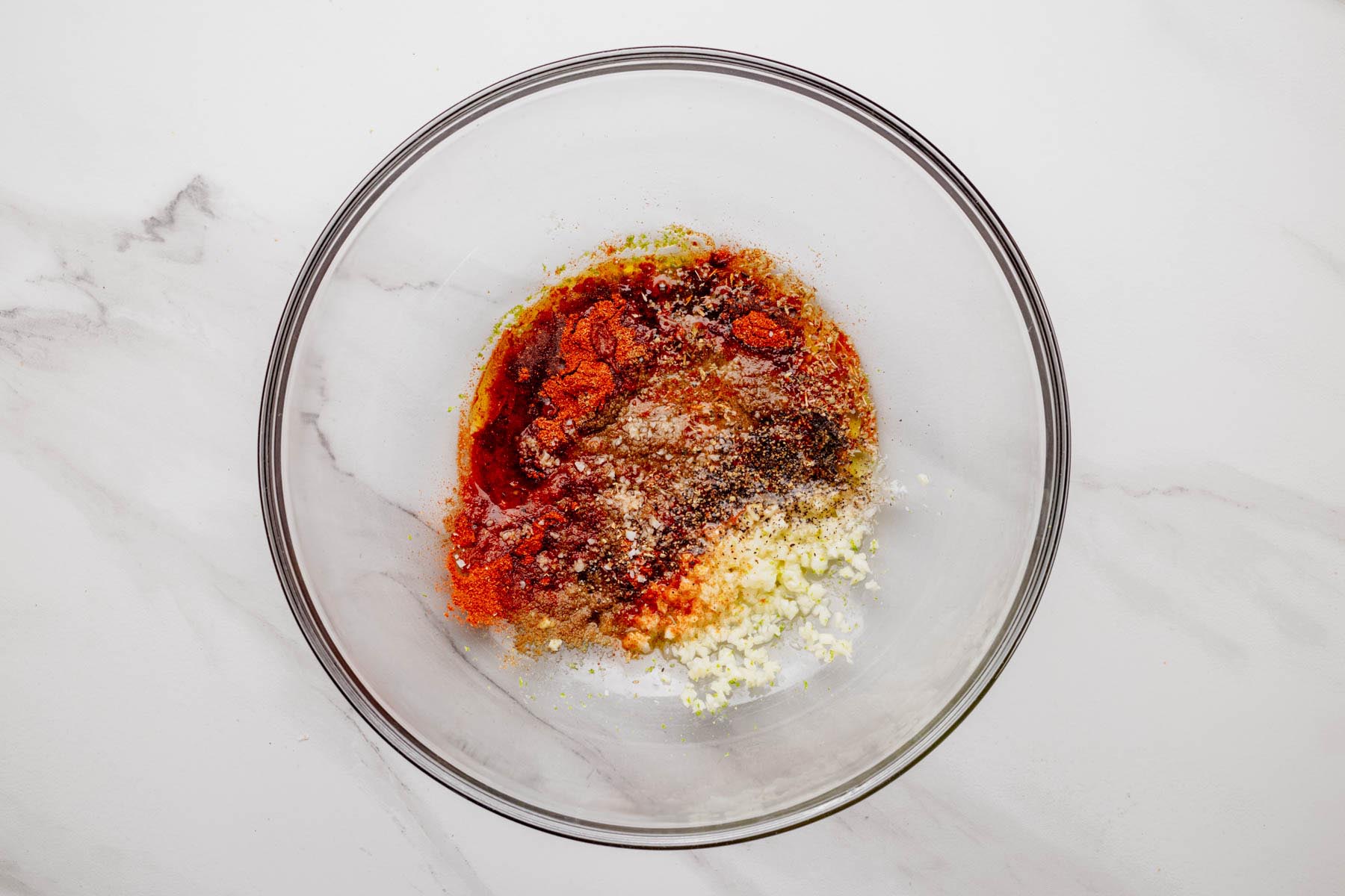 Glass bowl with unmixed marinade ingredients—olive oil, lime juice and zest, minced garlic, cumin, smoked paprika, oregano, salt, and pepper—ready to whisk.