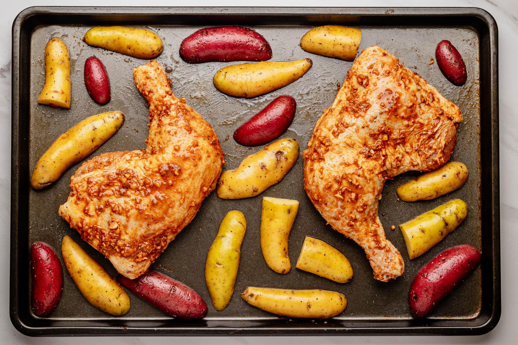 Marinated chicken leg quarters arranged skin-side up on a sheet pan with seasoned fingerling potatoes spaced around them, ready to roast.
