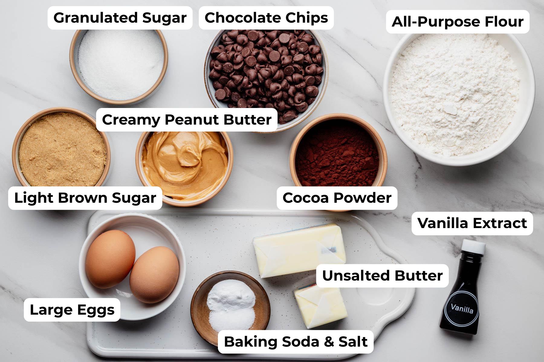Overhead view of all ingredients needed for chocolate chocolate chip peanut butter cookies arranged in bowls and labeled with text on a white marble surface.