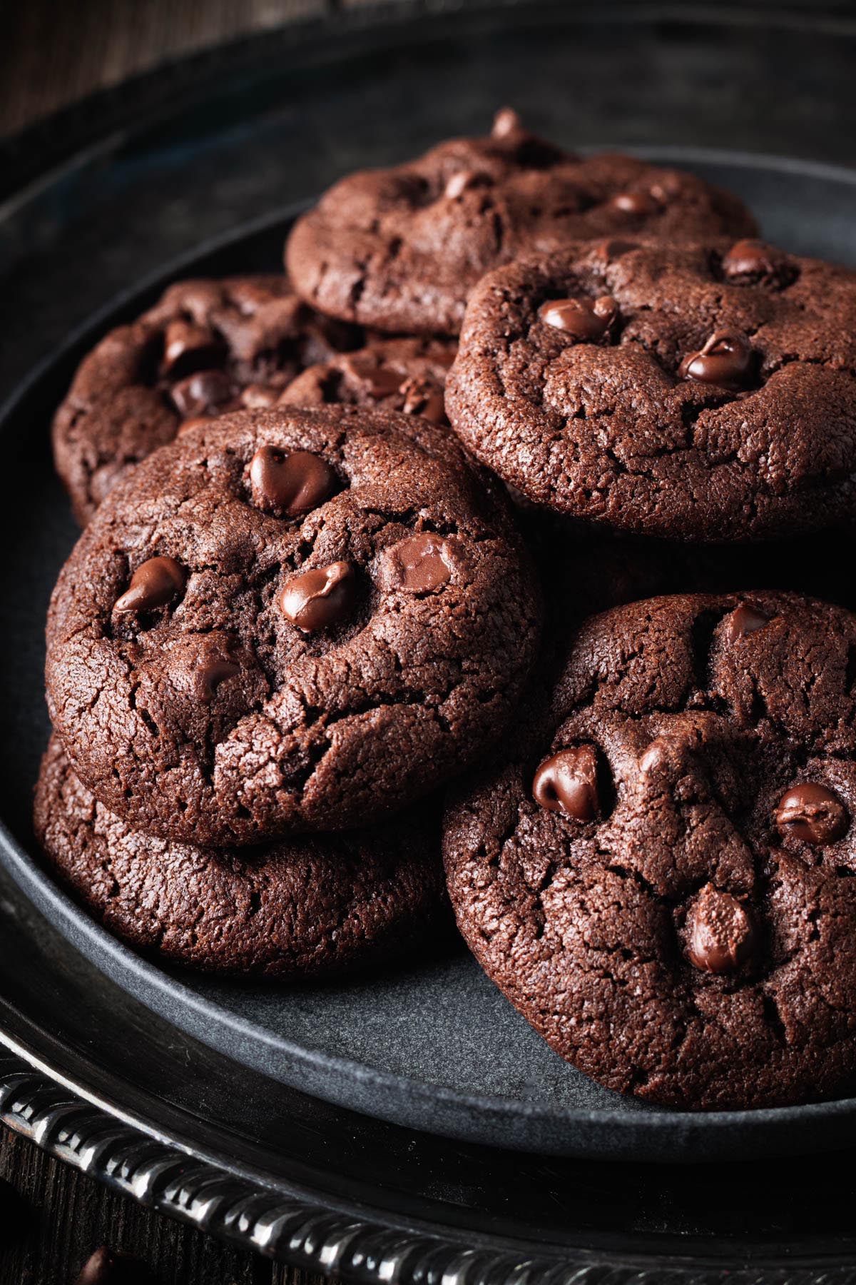 A close-up of chocolate peanut butter cookies piled on a dark plate, emphasizing their fudgy centers and chocolate chips.
