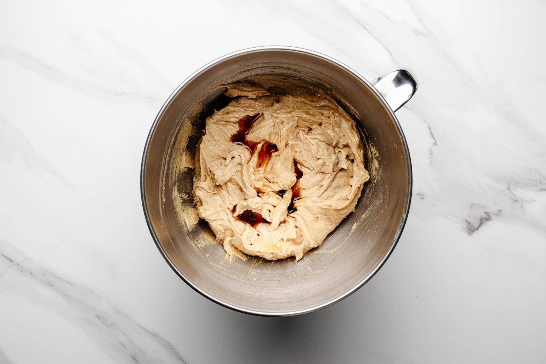 Smooth, creamed cookie dough in a mixer bowl with vanilla extract poured over the surface, ready to be mixed in.