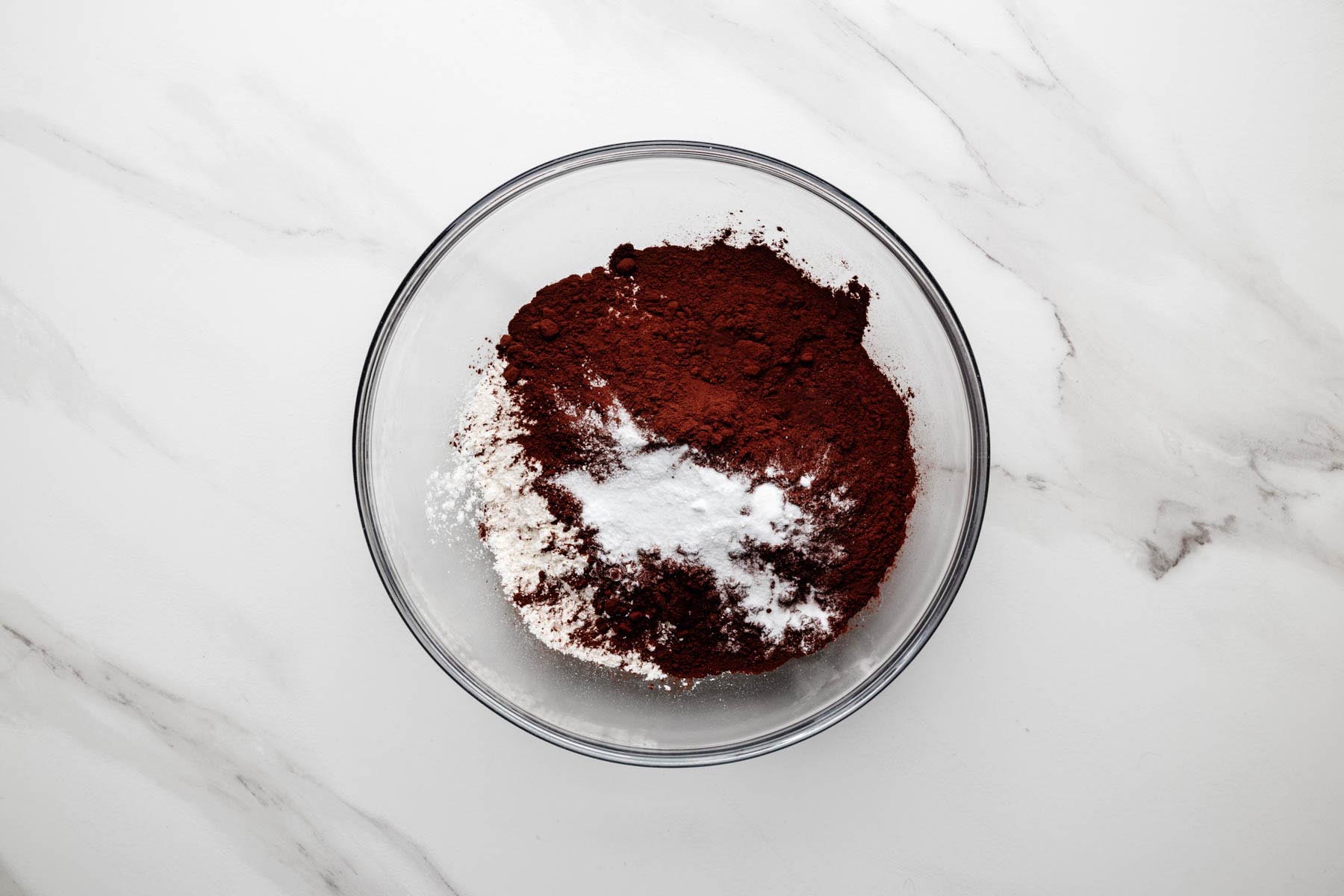 Dry ingredients (flour, cocoa powder, baking soda, and salt) in a glass mixing bowl on a white marble surface.