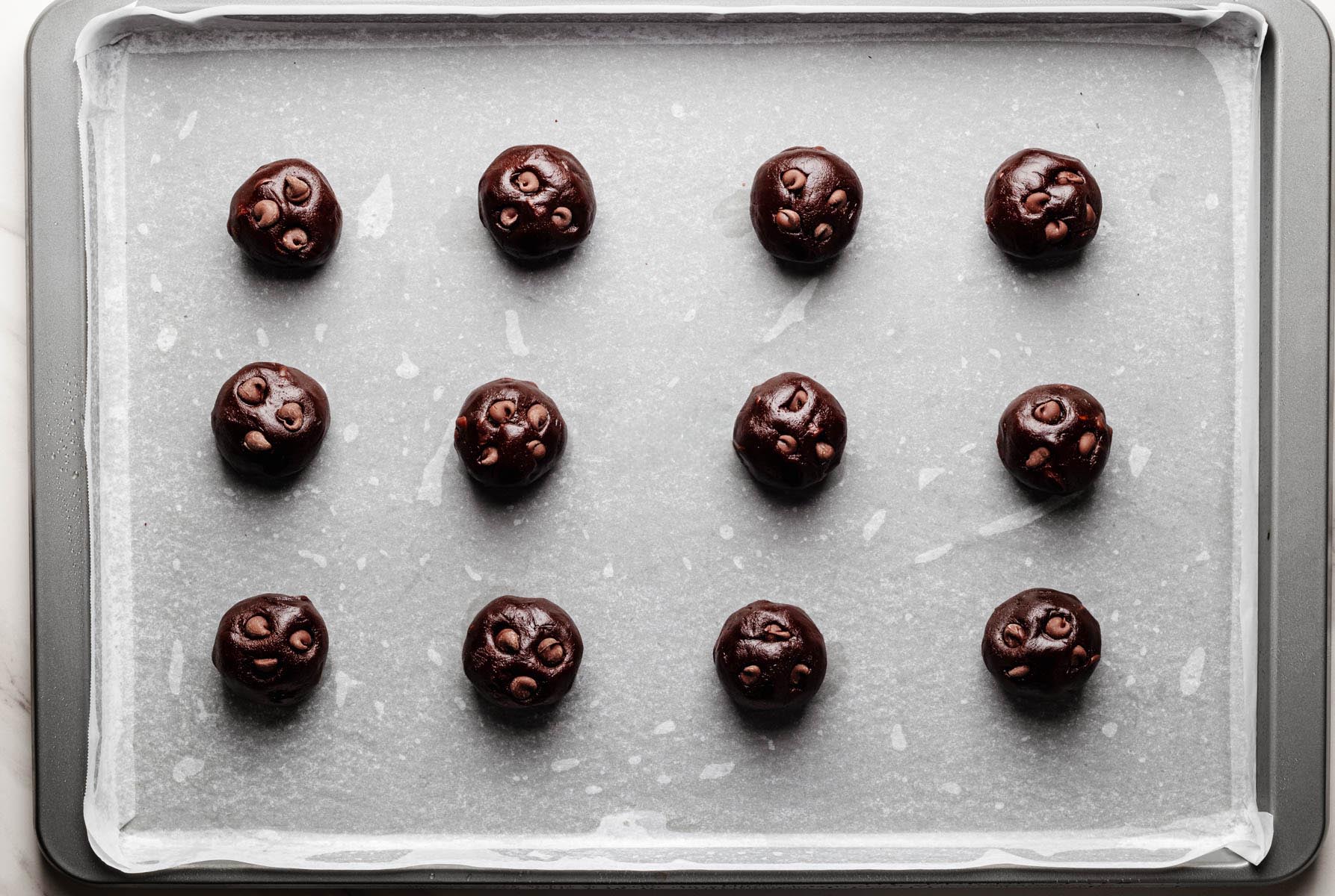 Portioned chocolate peanut butter cookie dough balls with visible chocolate chips arranged on a parchment-lined baking sheet.