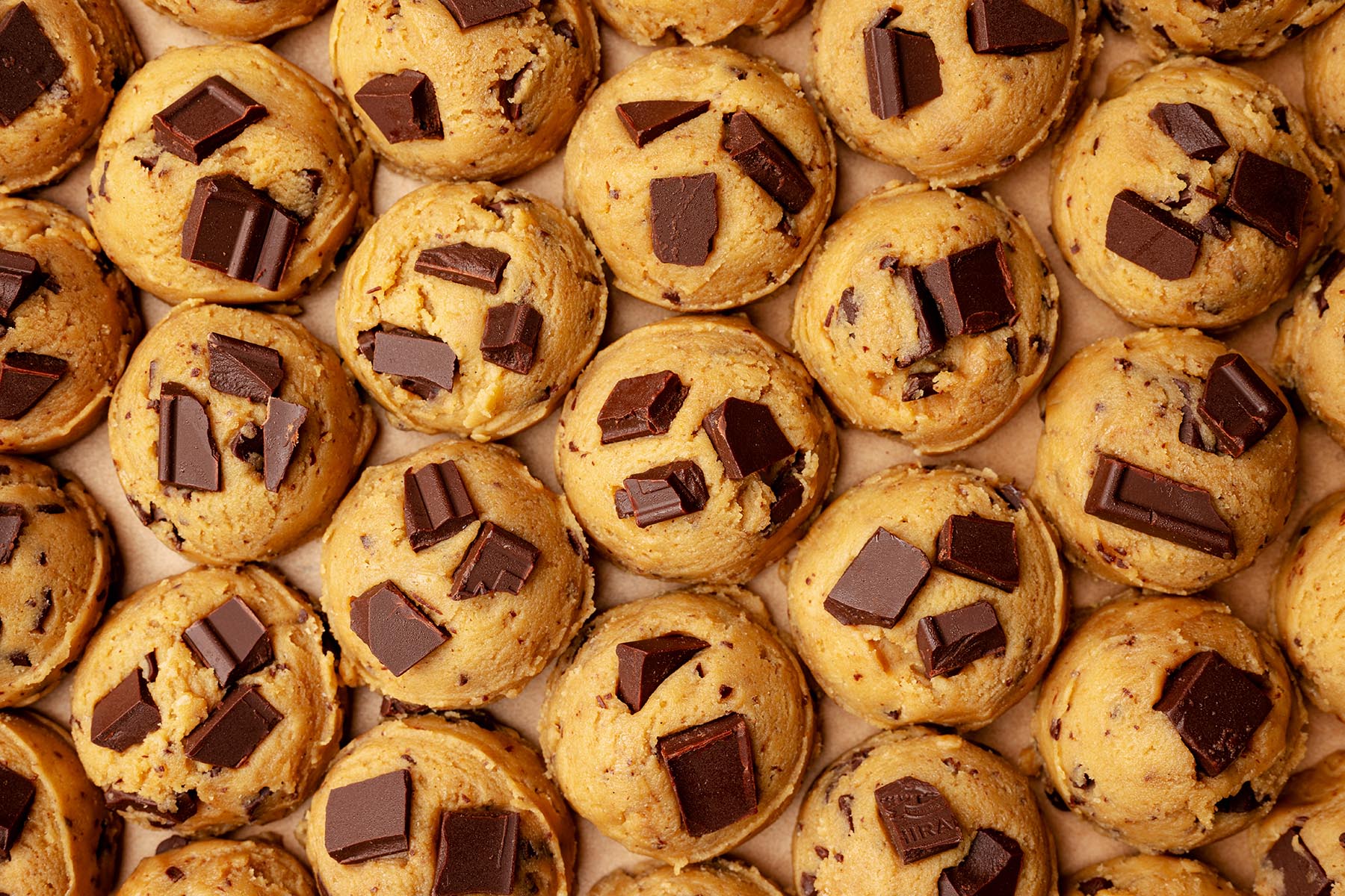 Rows of rounded cookie dough balls on a parchment-lined tray, each topped with extra chocolate chunks.