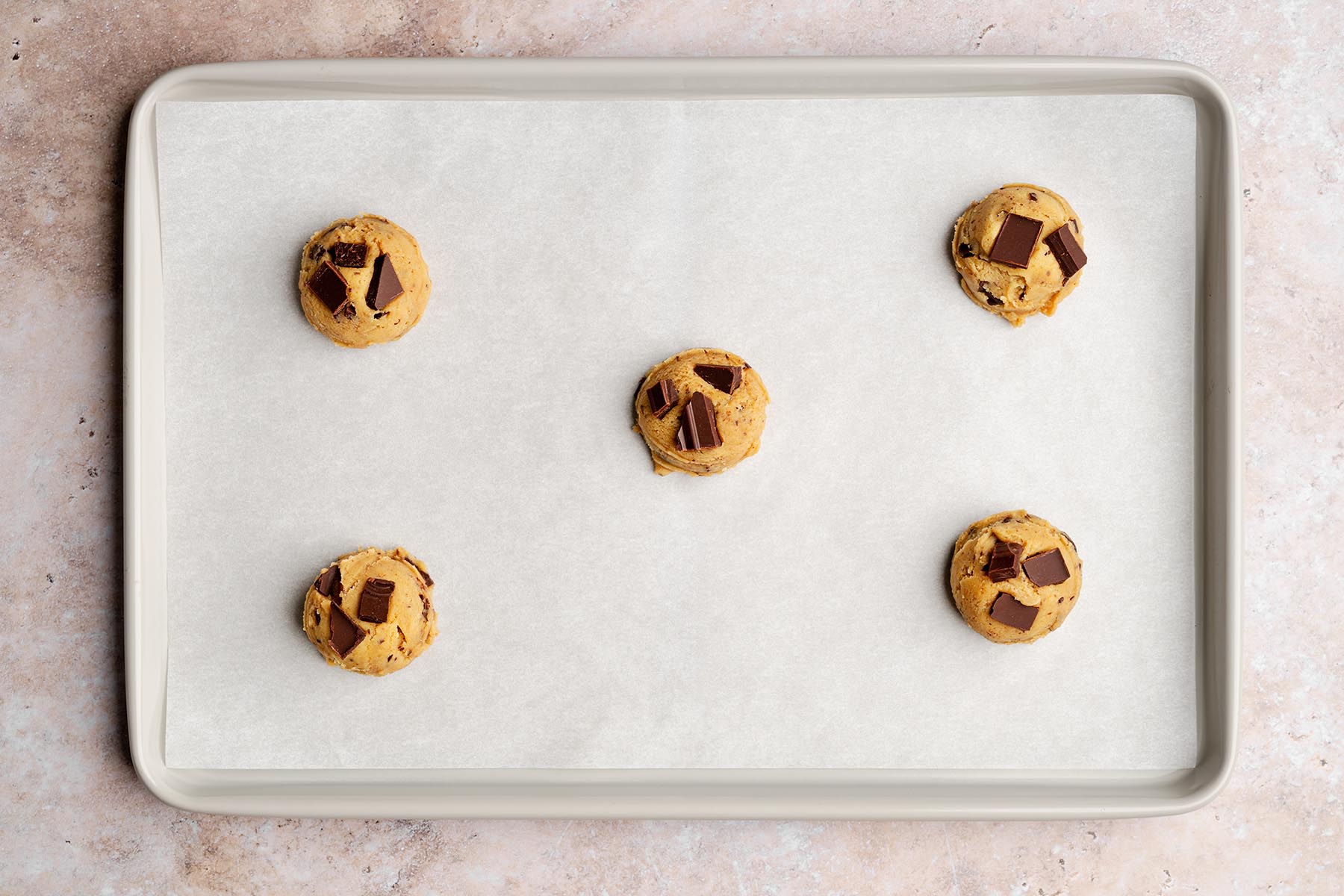 Five rounded chocolate chunk cookie dough balls arranged on a parchment-lined baking sheet, each topped with extra chocolate chunks before baking.
