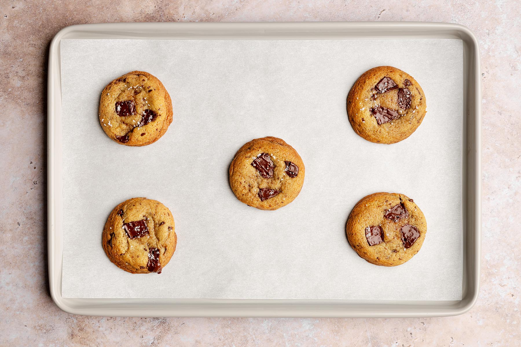 Five baked chocolate chunk cookies on a parchment-lined baking sheet, spaced apart with shiny melted chocolate pieces on top.