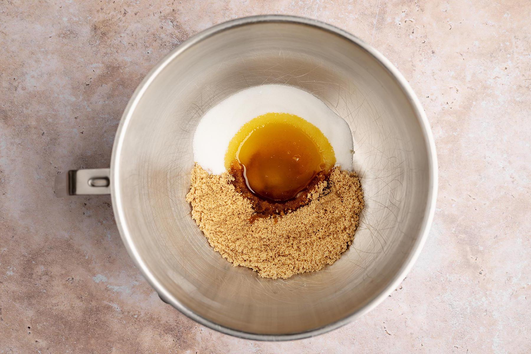 Overhead view of a metal mixing bowl with granulated sugar, brown sugar, and a pool of cooled browned butter in the center.