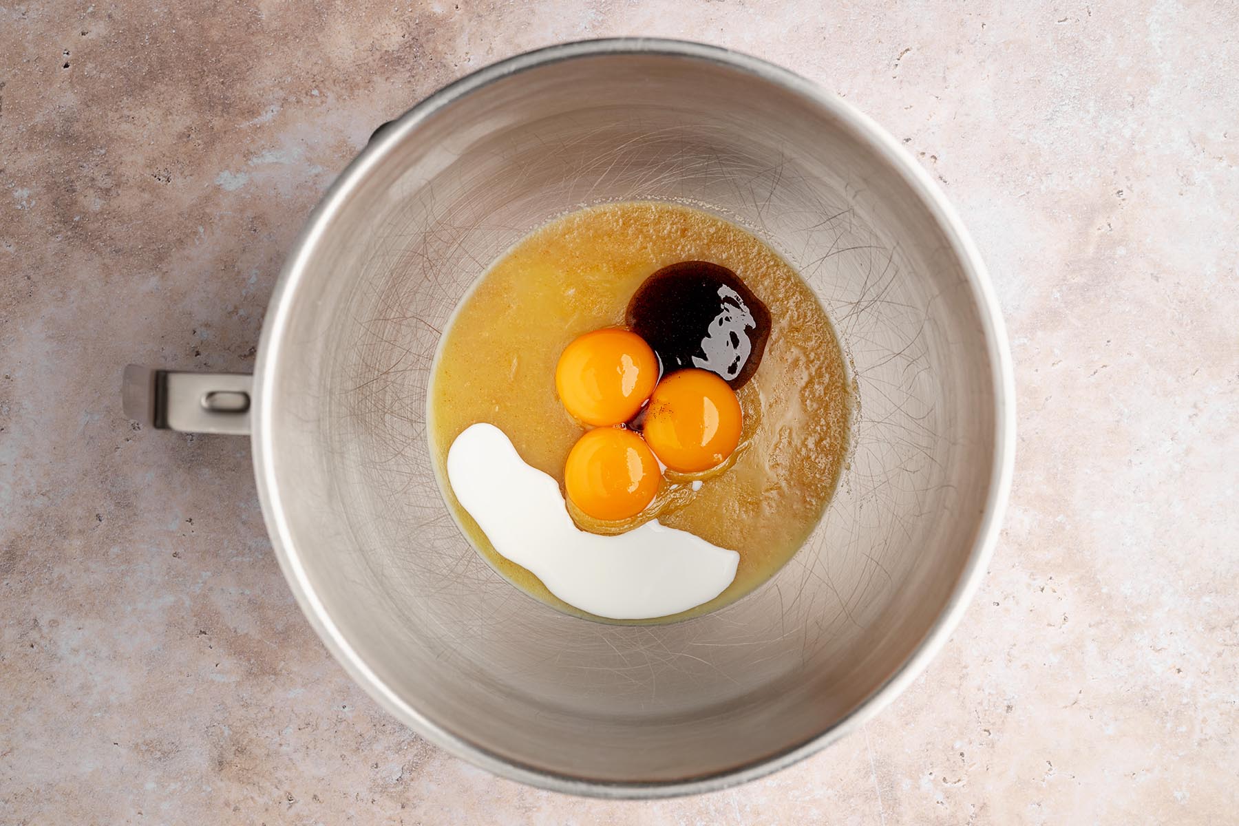Metal mixing bowl with browned butter mixture topped with three egg yolks, vanilla, and a splash of milk before mixing.