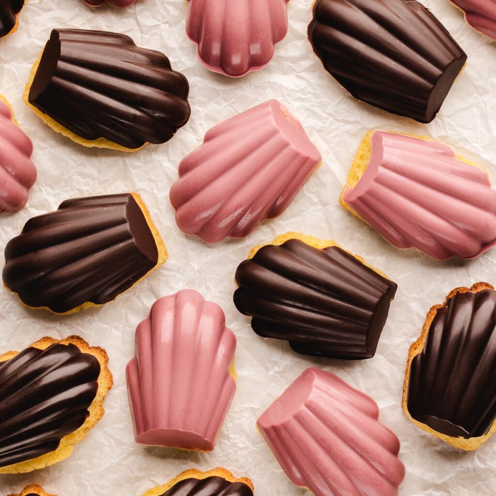 Overhead of chocolate-dipped and ruby-coated madeleines arranged on crinkled parchment, showing the scalloped shells.