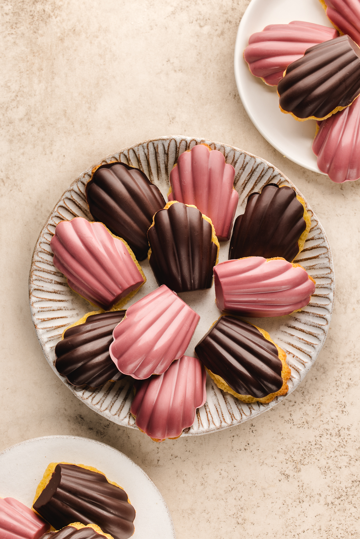 Assorted ruby and dark chocolate madeleines arranged on a ceramic serving plate with a few on side plates.