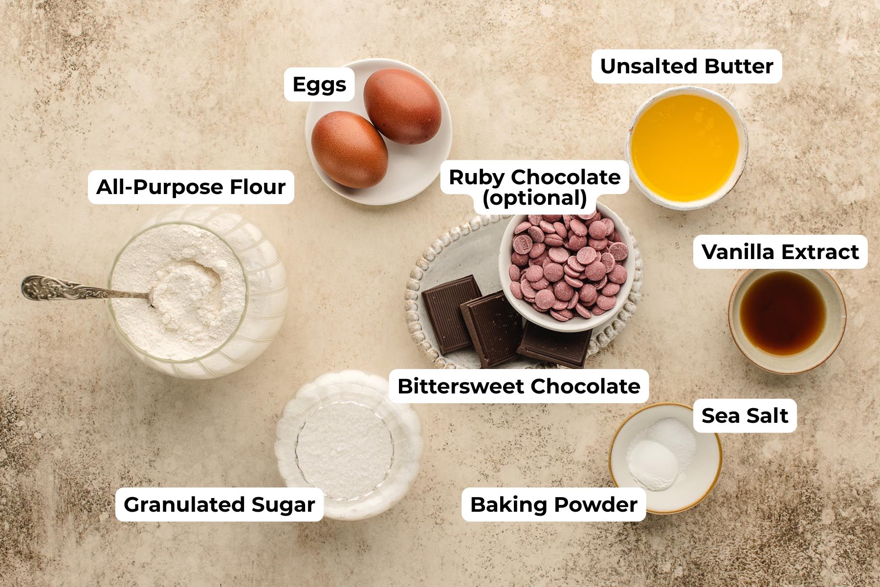 Overhead photo of labeled ingredients for Chocolate Dipped Madeleines: all-purpose flour, granulated sugar, eggs, melted unsalted butter, vanilla extract, sea salt, baking powder, bittersweet chocolate, and optional ruby chocolate callets on a neutral stone surface.
