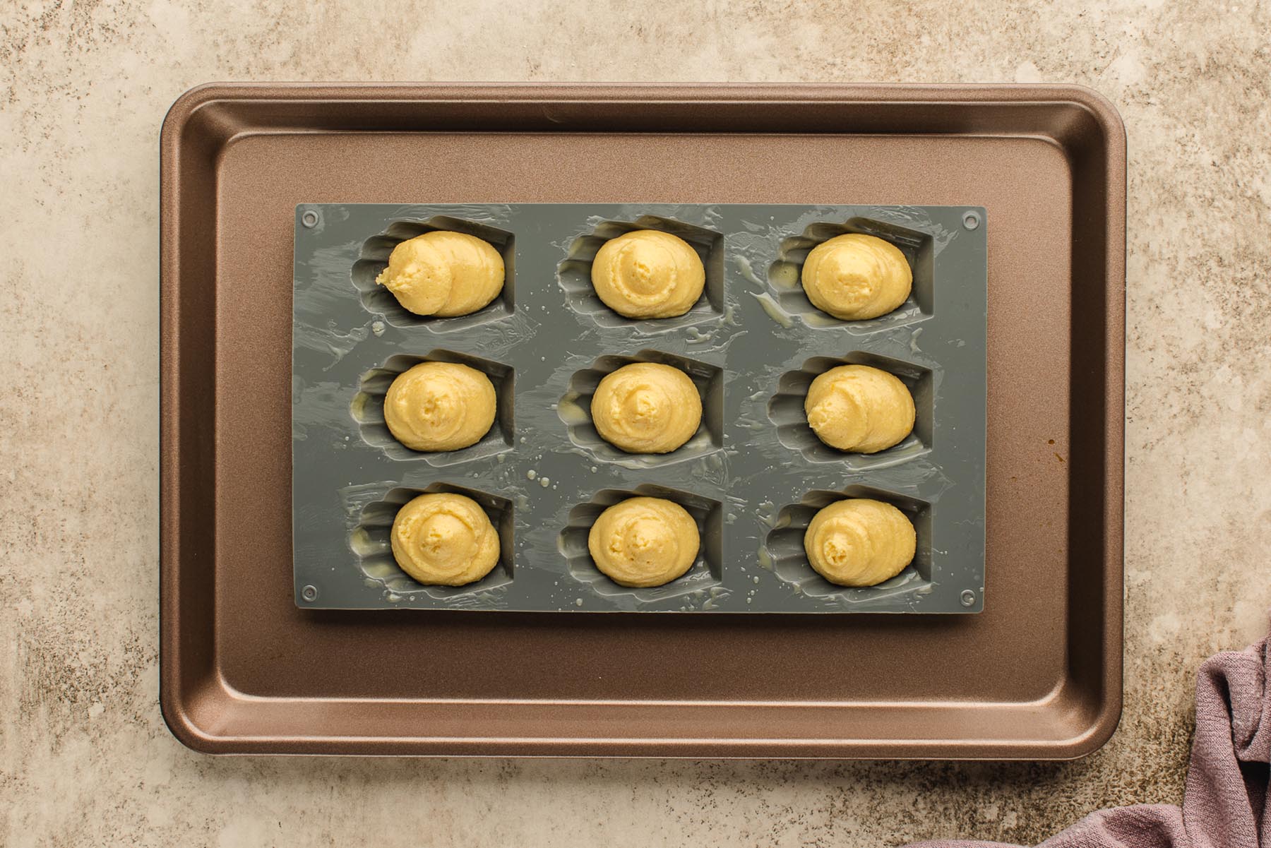 Mounds of batter piped into a greased silicone madeleine pan set on a sheet tray.