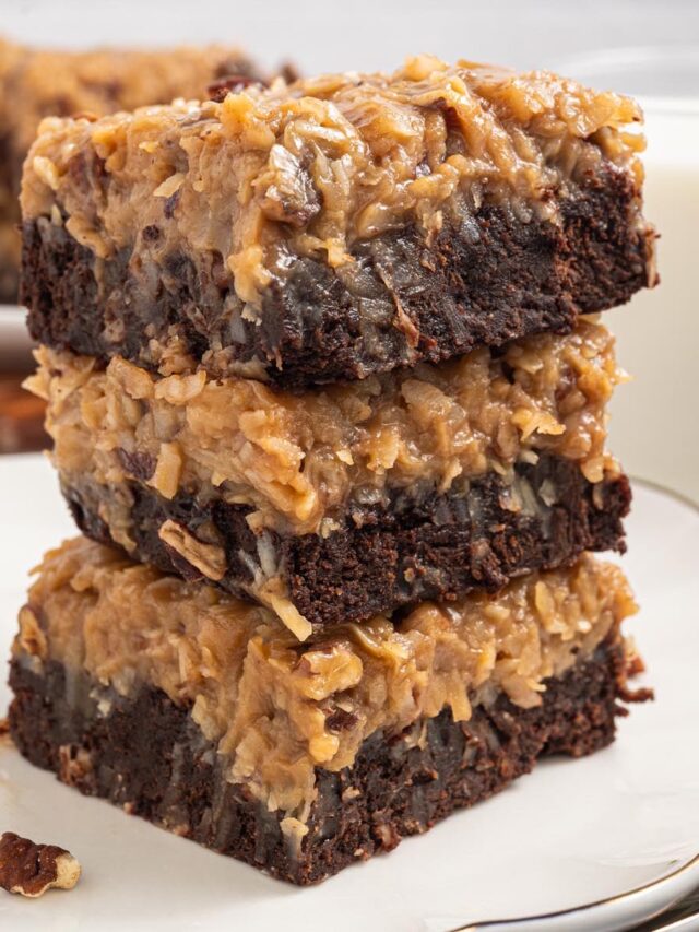 Close-up stack of three German chocolate brownies with coconut-pecan frosting on a white plate, fudgy edges and crumb visible; glass of milk behind.