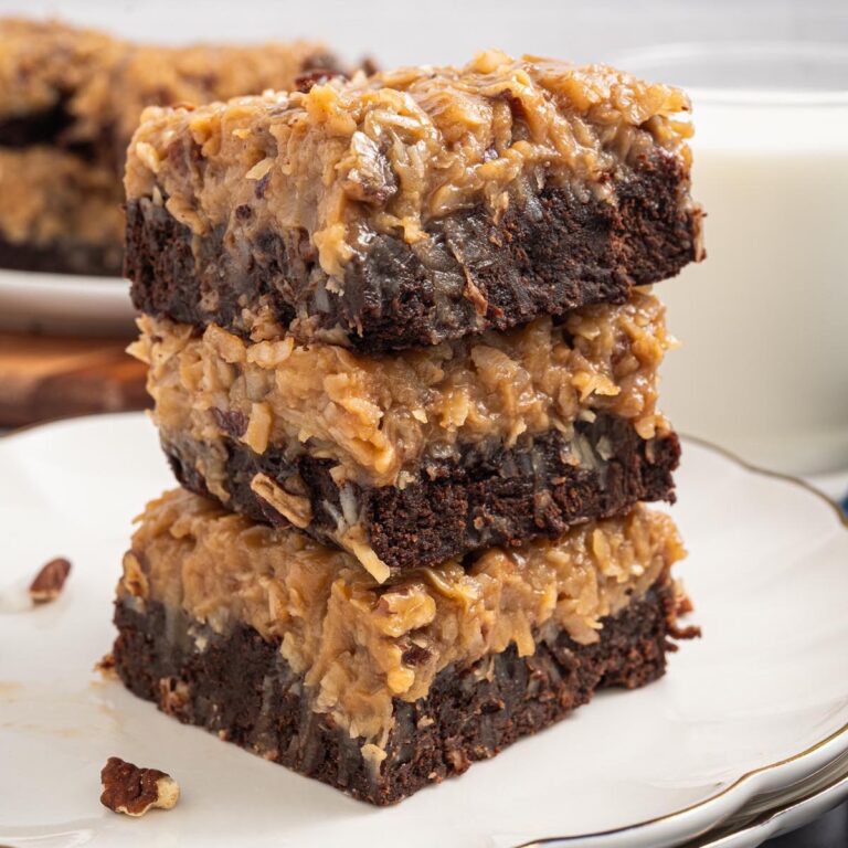 Close-up stack of three German chocolate brownies with coconut-pecan frosting on a white plate, fudgy edges and crumb visible; glass of milk behind.
