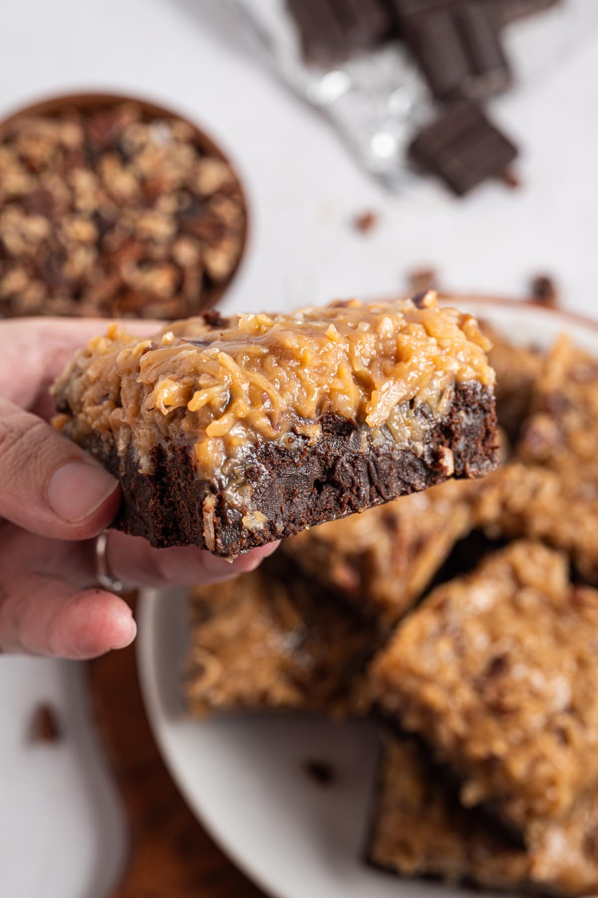 Hand holding a German chocolate brownie to show the fudgy interior and gooey coconut-pecan frosting; plate of brownies and chocolate/pecans blurred in the background.