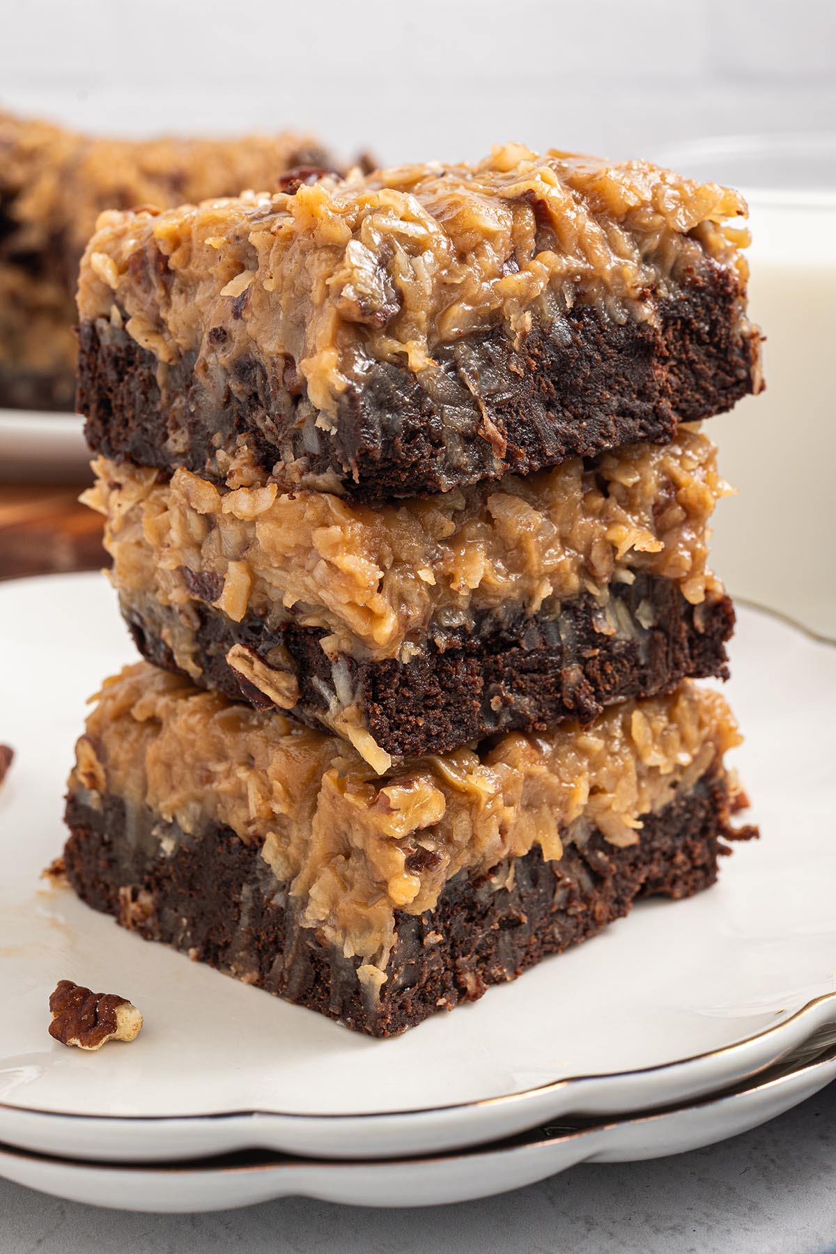 Three German chocolate brownies stacked on a white plate, showing a fudgy chocolate base and thick coconut-pecan frosting; milk in the background.