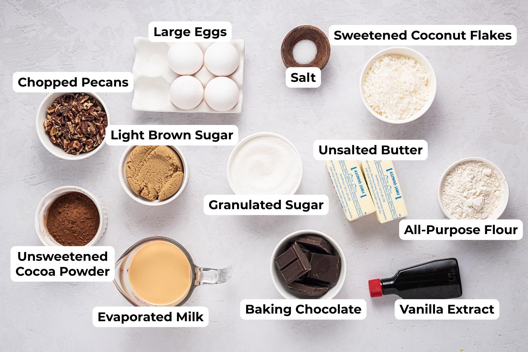 Overhead shot of all ingredients for German Chocolate Brownies arranged on a light surface with labels—eggs, sugars, butter, flour, cocoa powder, baking chocolate, evaporated milk, vanilla, coconut flakes, pecans, and salt.