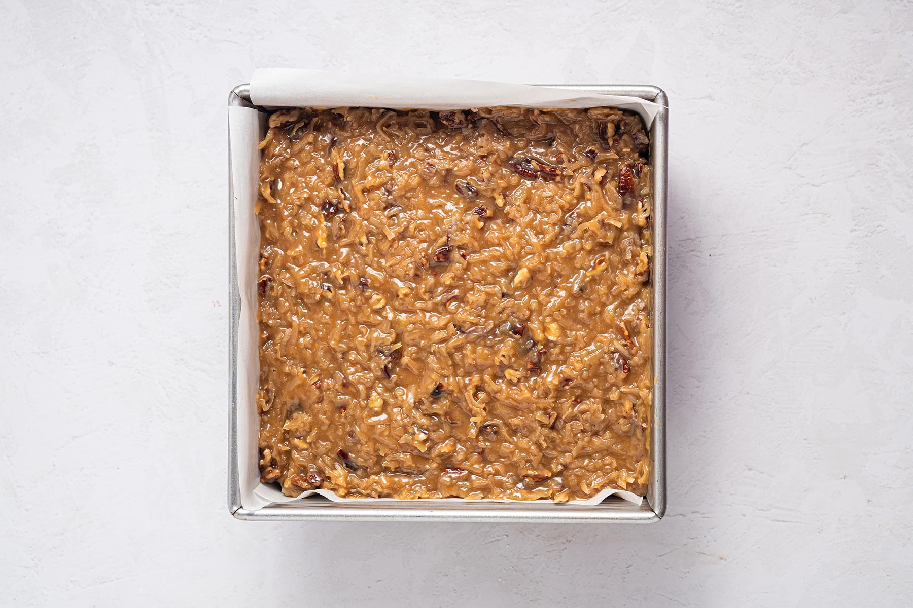 Brownies topped edge-to-edge with coconut-pecan frosting in the parchment-lined pan, ready to chill and slice.