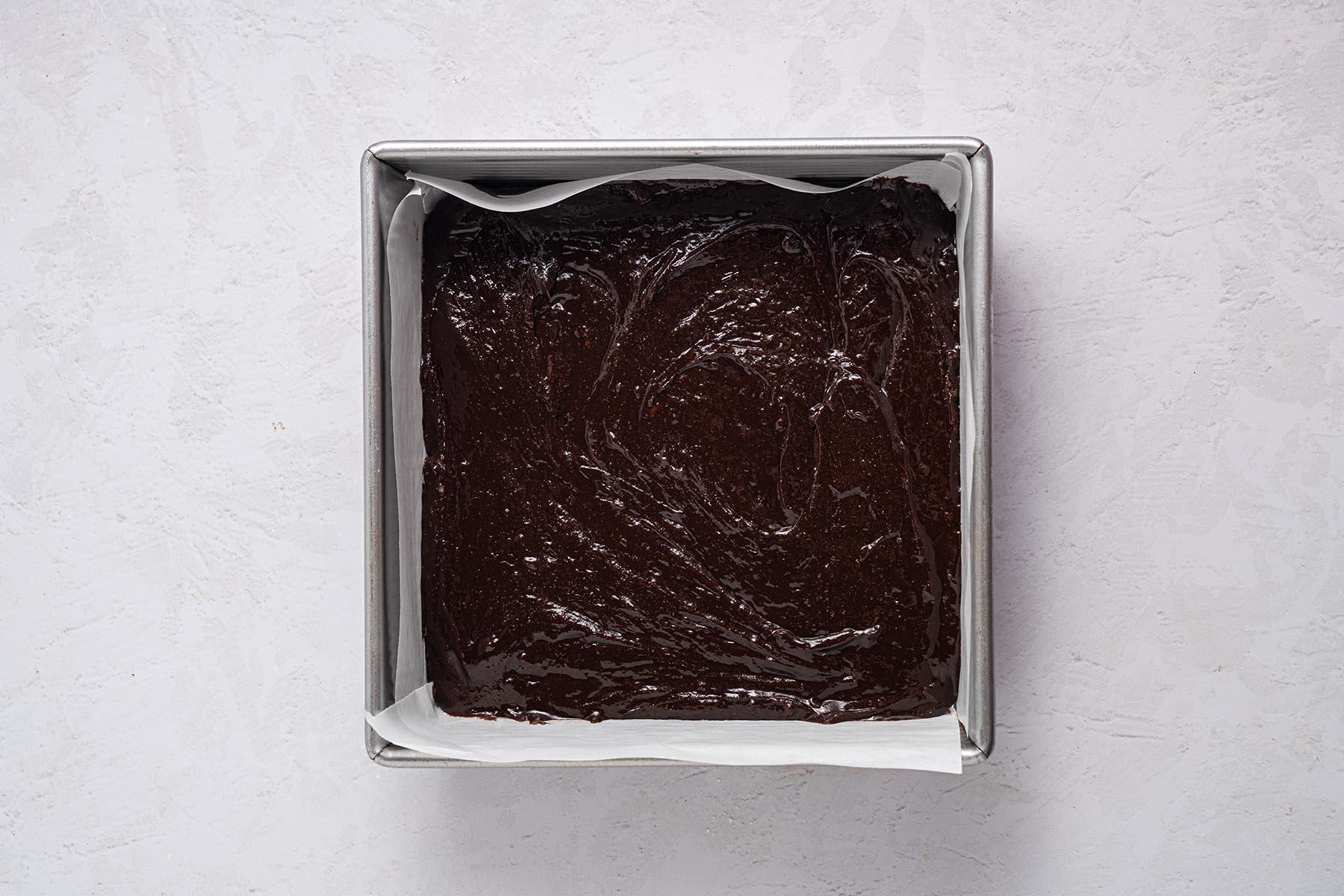 Brownie batter spread in a parchment-lined 8×8 pan with swirled top, ready to bake.
