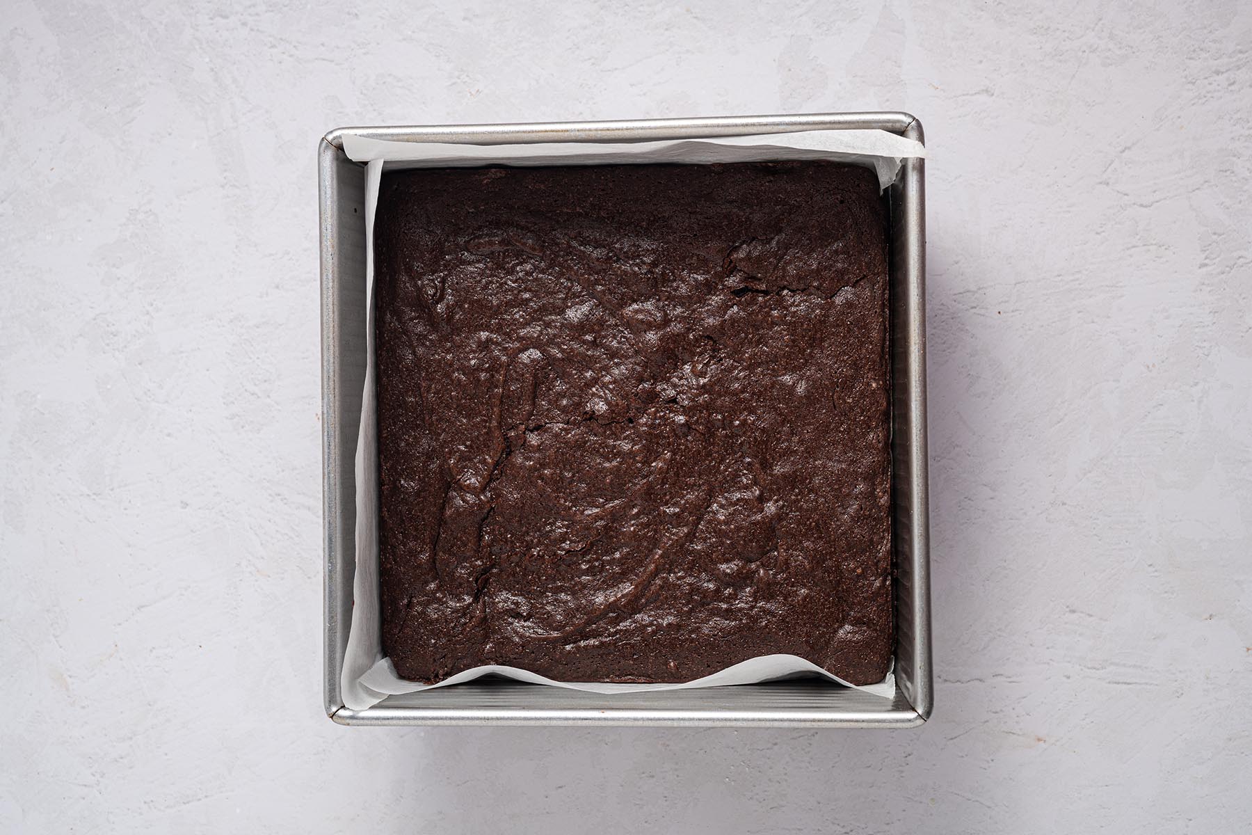 Fully baked brownie slab in a parchment-lined 8×8 pan, edges set and surface matte, cooling before frosting.