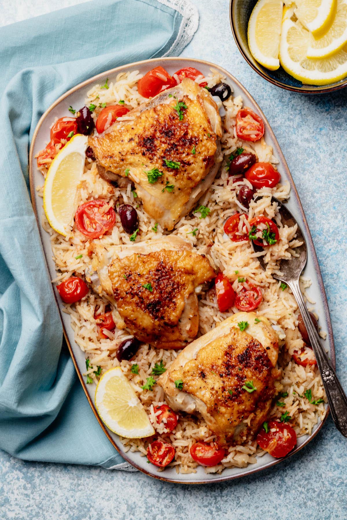 Platter of Greek chicken and rice with cherry tomatoes, olives, and lemon wedges, overhead view.