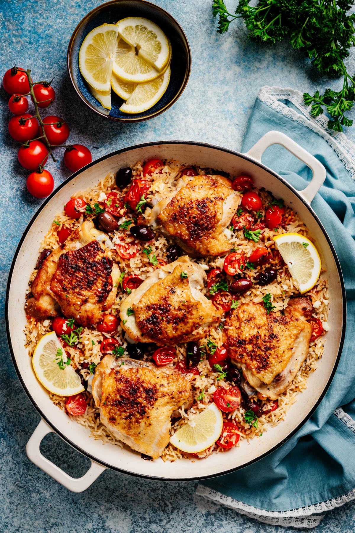 Overhead of Greek chicken on herbed rice with tomatoes and olives; lemon slices and napkin at the side.