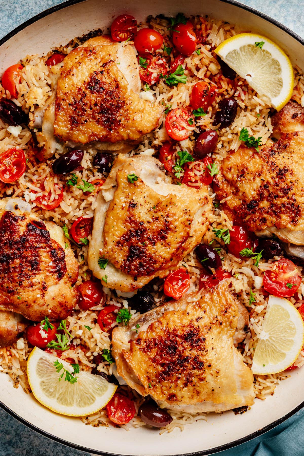 Close-up of crispy chicken thighs nestled in tomato-olive rice with parsley and lemon.