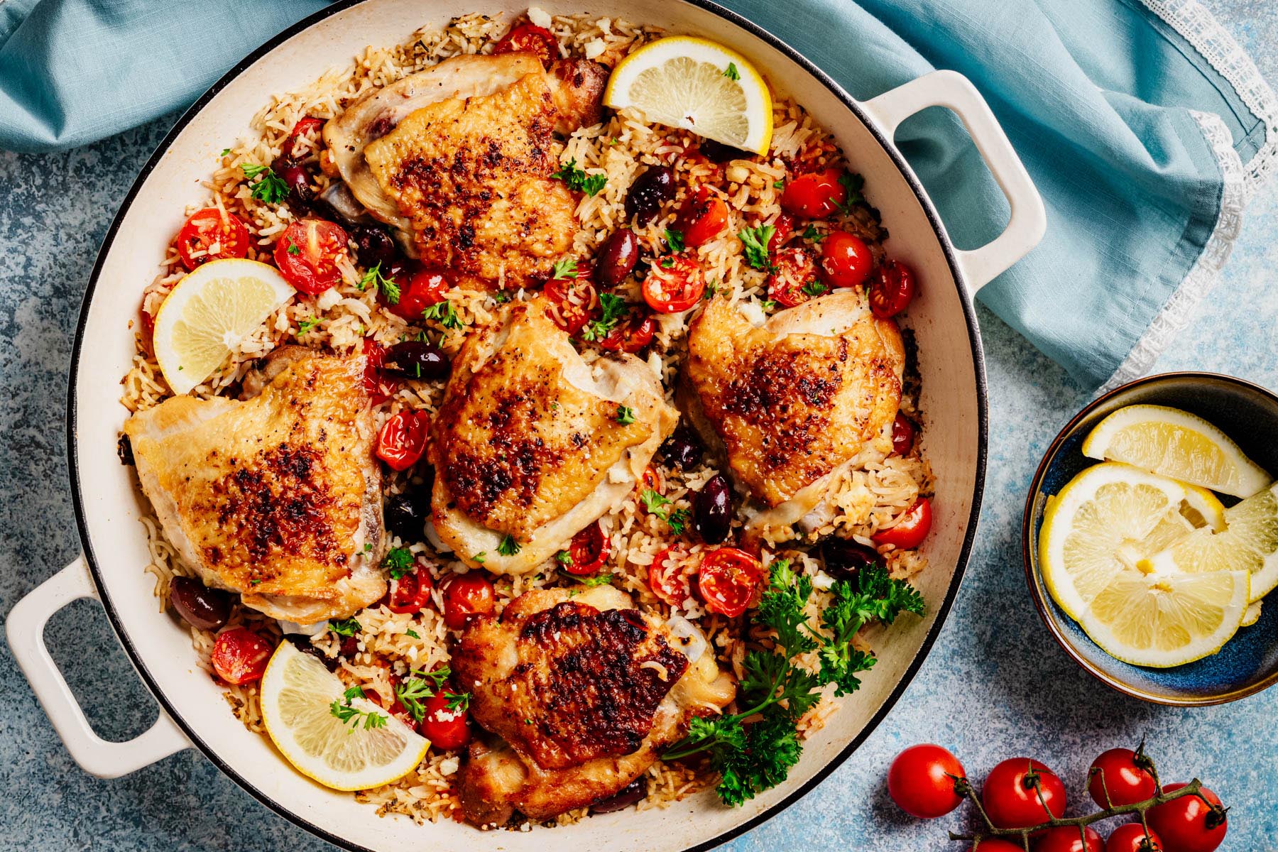 Family-style skillet of Greek chicken and rice with tomatoes, olives, parsley, and lemon wedges, ready to serve.