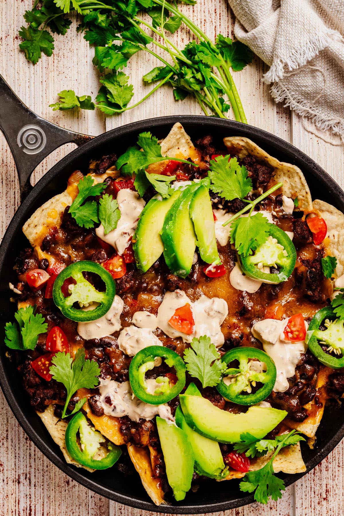 Close-up of loaded beef nachos topped with melted cheese, sliced avocado, jalapeños, fresh cilantro, diced tomatoes, and sour cream, served in a black skillet.