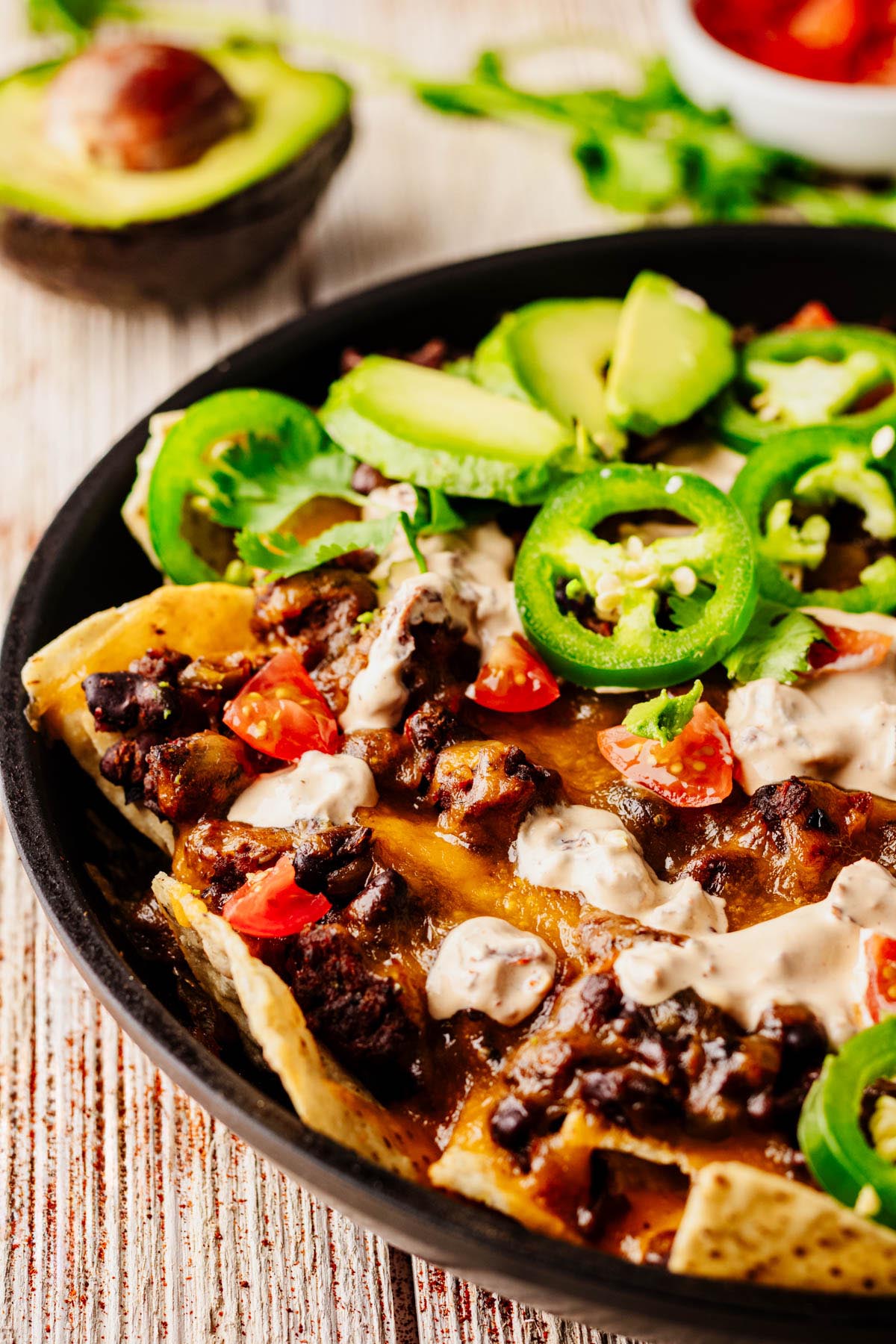Close-up of loaded beef nachos with melted cheese, ground beef, diced tomatoes, jalapeños, sour cream, and sliced avocado in a black skillet, with an avocado and fresh cilantro in the background.