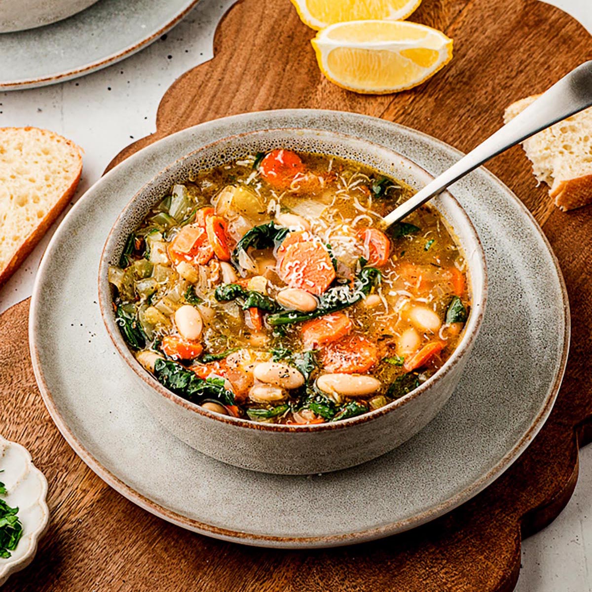 Close-up of a bowl of Tuscan white bean soup topped with grated Parmesan, showing carrots, white beans, spinach, and celery in a golden broth on a gray plate.