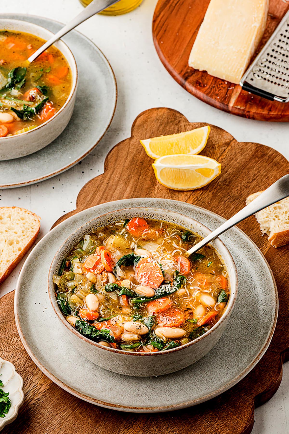 Bowl of Tuscan white bean soup with carrots, spinach, and cannellini beans on a gray plate and wood board, with lemon wedges, bread, and a second bowl in the background.