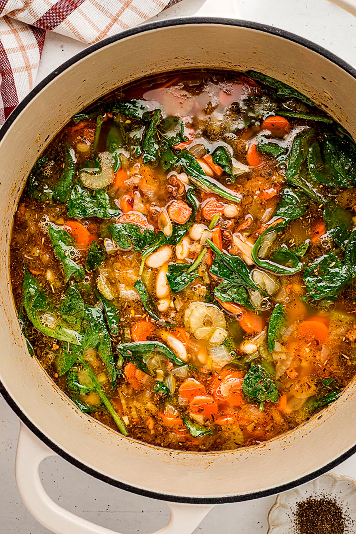 Overhead shot of Tuscan white bean soup simmering in a Dutch oven, with visible spinach, carrots, celery, onions, and white beans in a herbed broth.