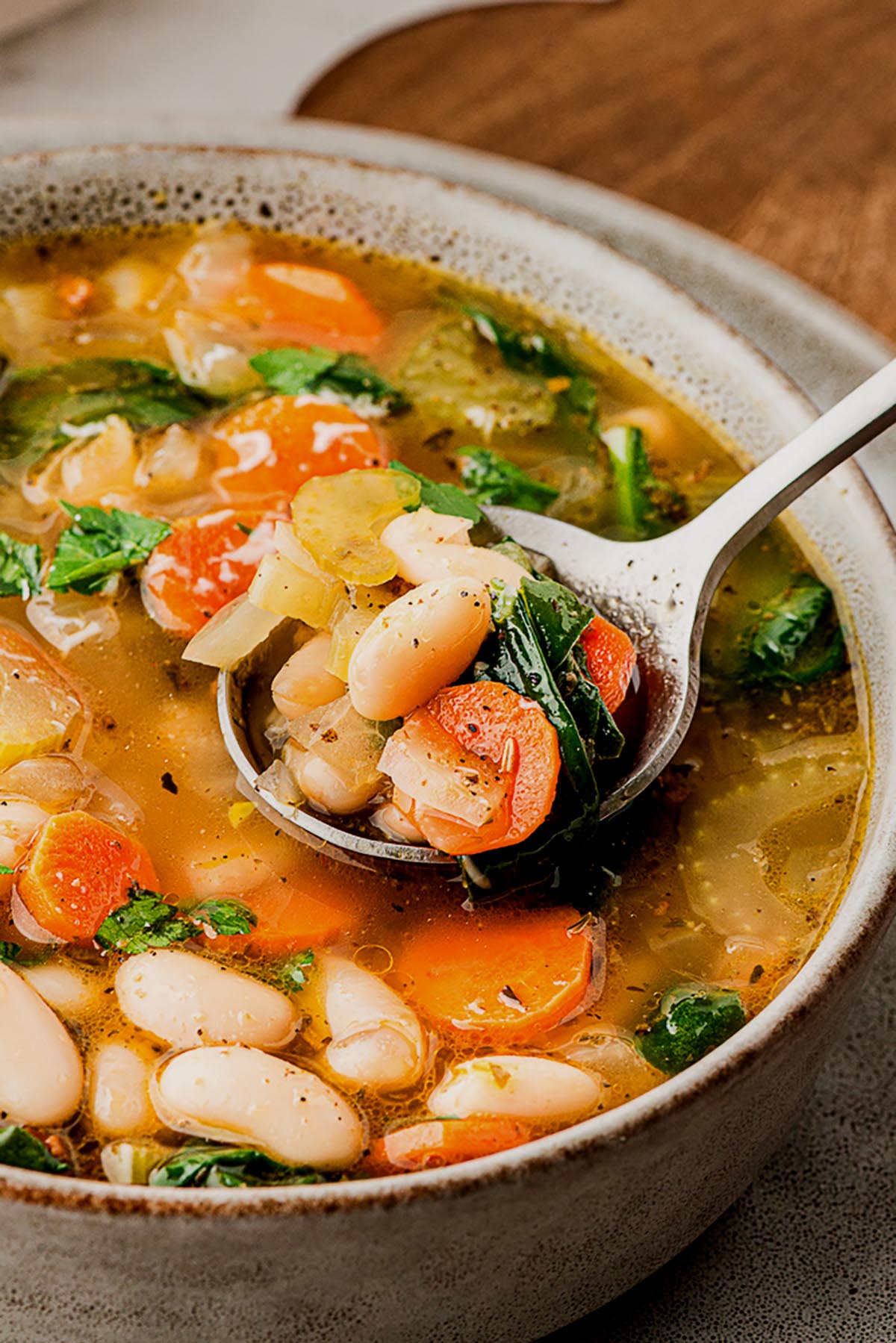 Close-up of a spoonful of Tuscan white bean soup lifted from a bowl, showing white beans, carrots, celery, spinach, and fresh herbs in a light broth.