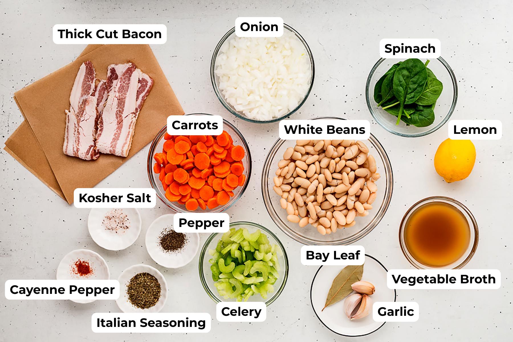 Overhead view of labeled ingredients for Tuscan white bean soup, including thick-cut bacon, diced onion, sliced carrots, chopped celery, white beans, baby spinach, lemon, vegetable broth, garlic, bay leaves, kosher salt, black pepper, cayenne pepper, and Italian seasoning arranged in small bowls on a light surface.