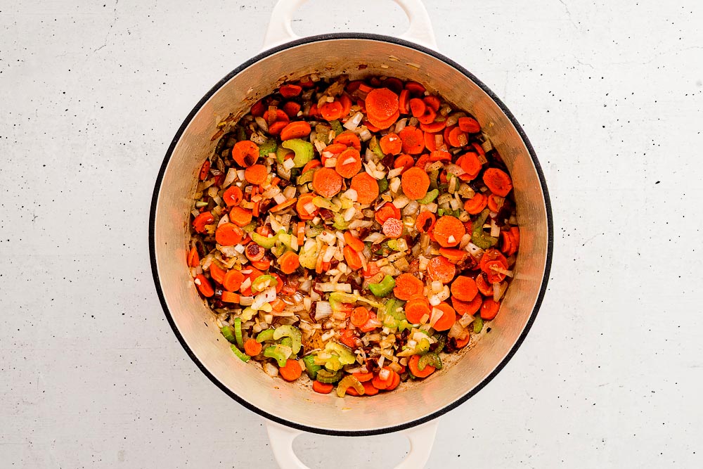 Overhead view of diced carrots, celery, and onions sautéing in a Dutch oven with the browned bacon bits.