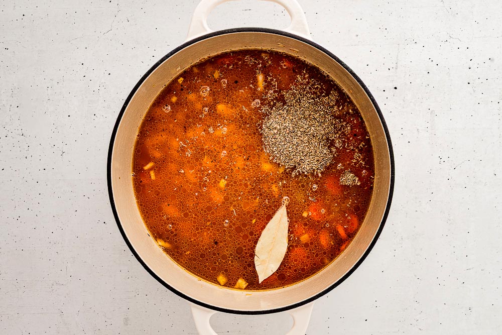 Overhead view of the soup base in a Dutch oven with broth, Italian seasoning, and a bay leaf added on top before simmering.