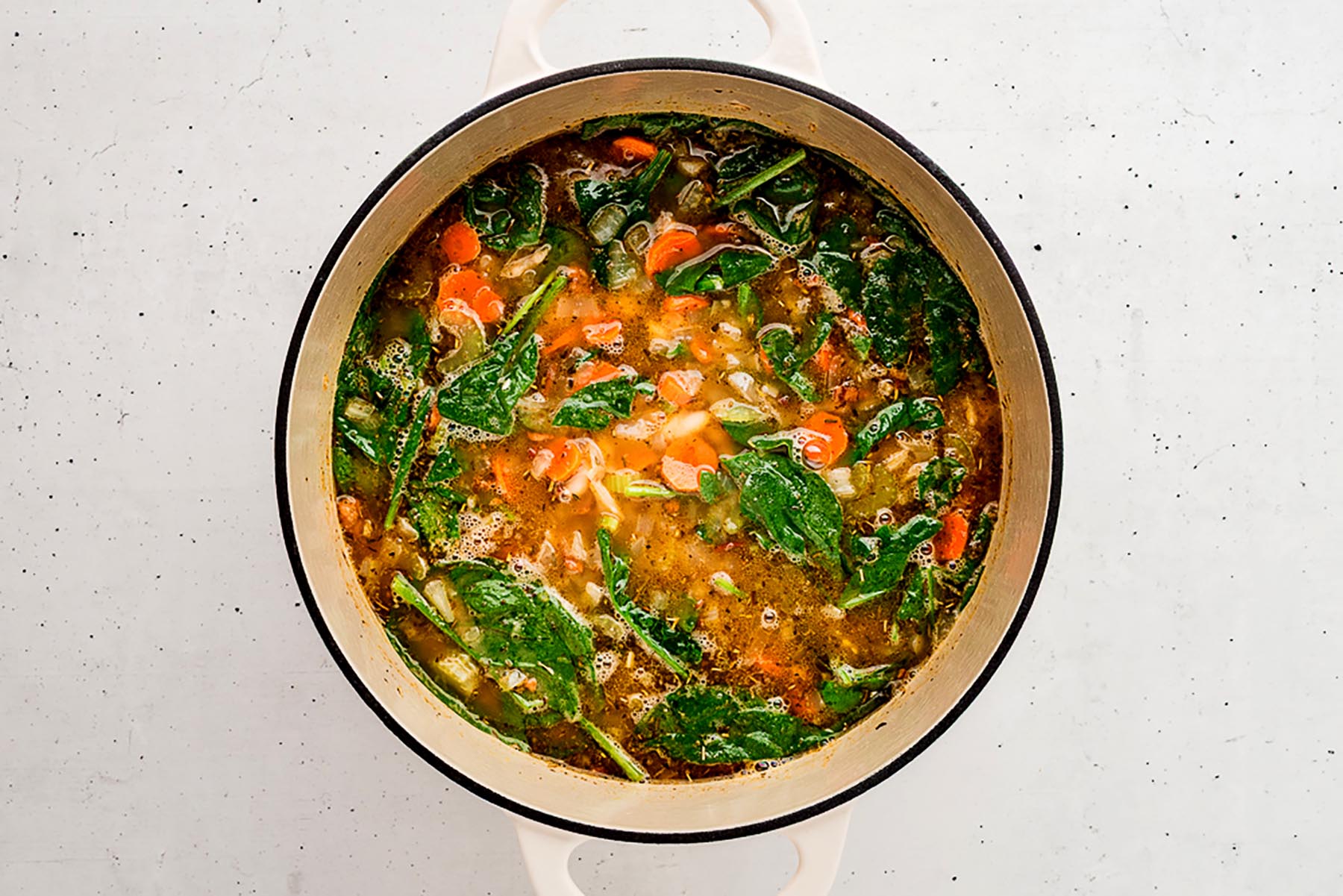 Overhead view of a Dutch oven filled with Tuscan white bean soup simmering on the stove, with carrots, celery, onions, and fresh spinach visible in the broth.