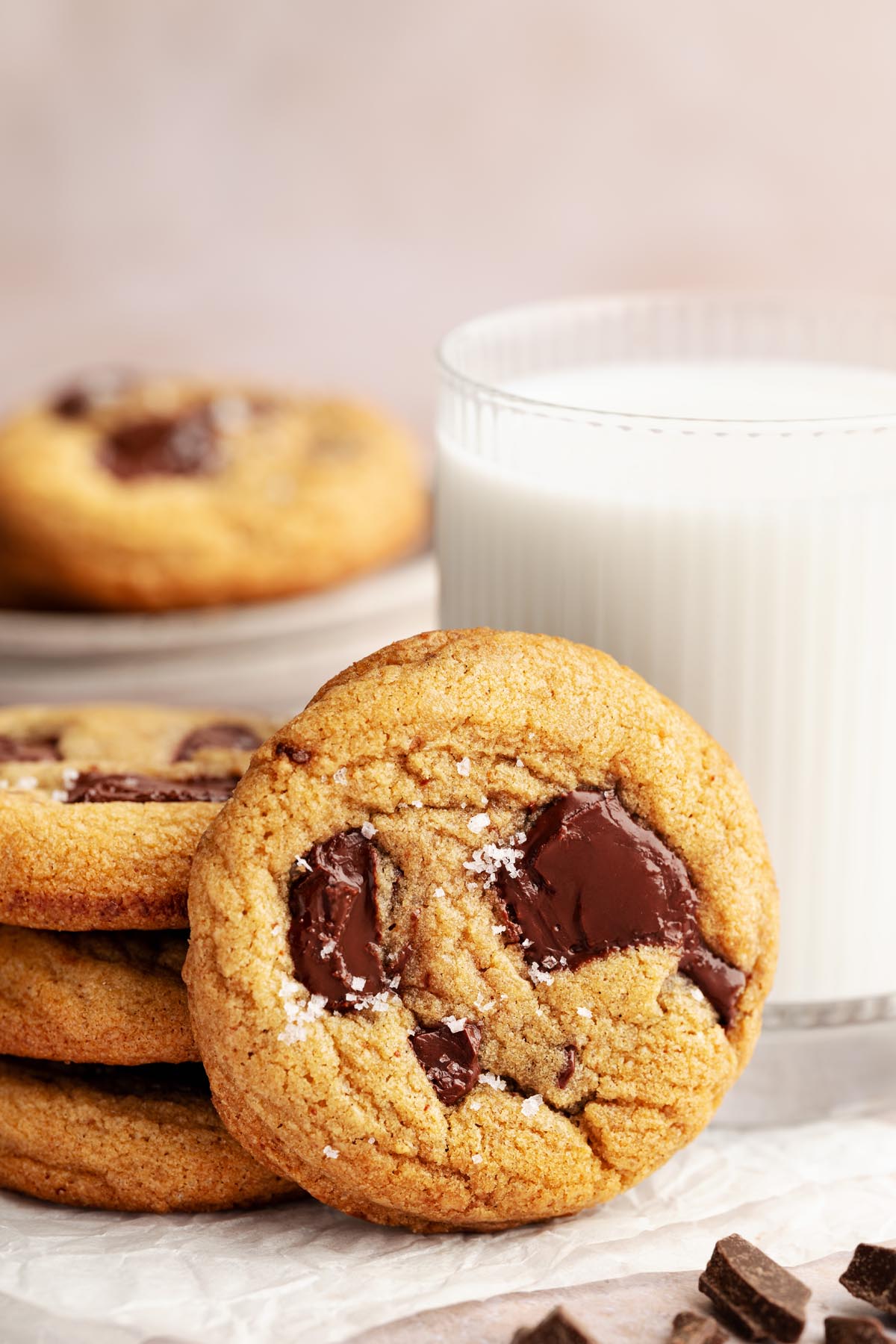 Chocolate chunk cookie leaning against a small stack of cookies with a glass of milk in the background and chocolate pieces scattered in front.